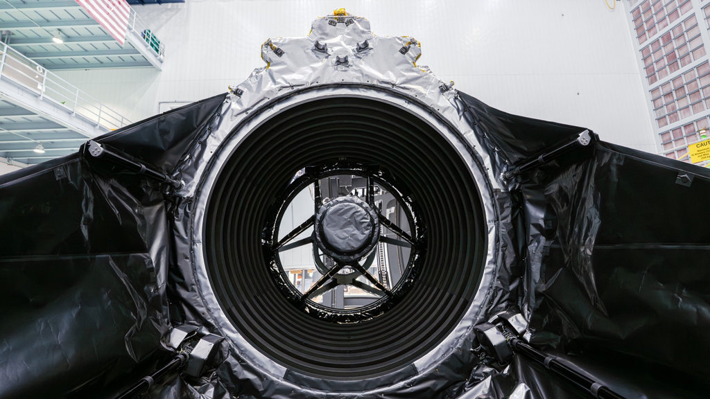 technicians in clean suits in a hangar next to a large rectangular spacecraft wrapped in metallic foil